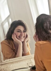 A cheerful woman smiles at her reflection in a vintage-style mirror, exuding positivity and warmth.